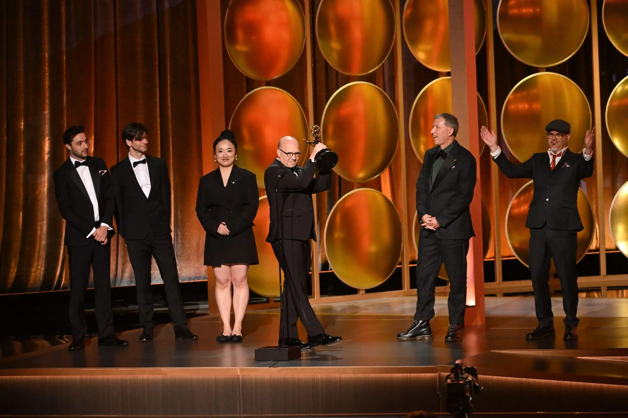 Five people on stage at awards ceremony, man holding trophy, orange and gold background