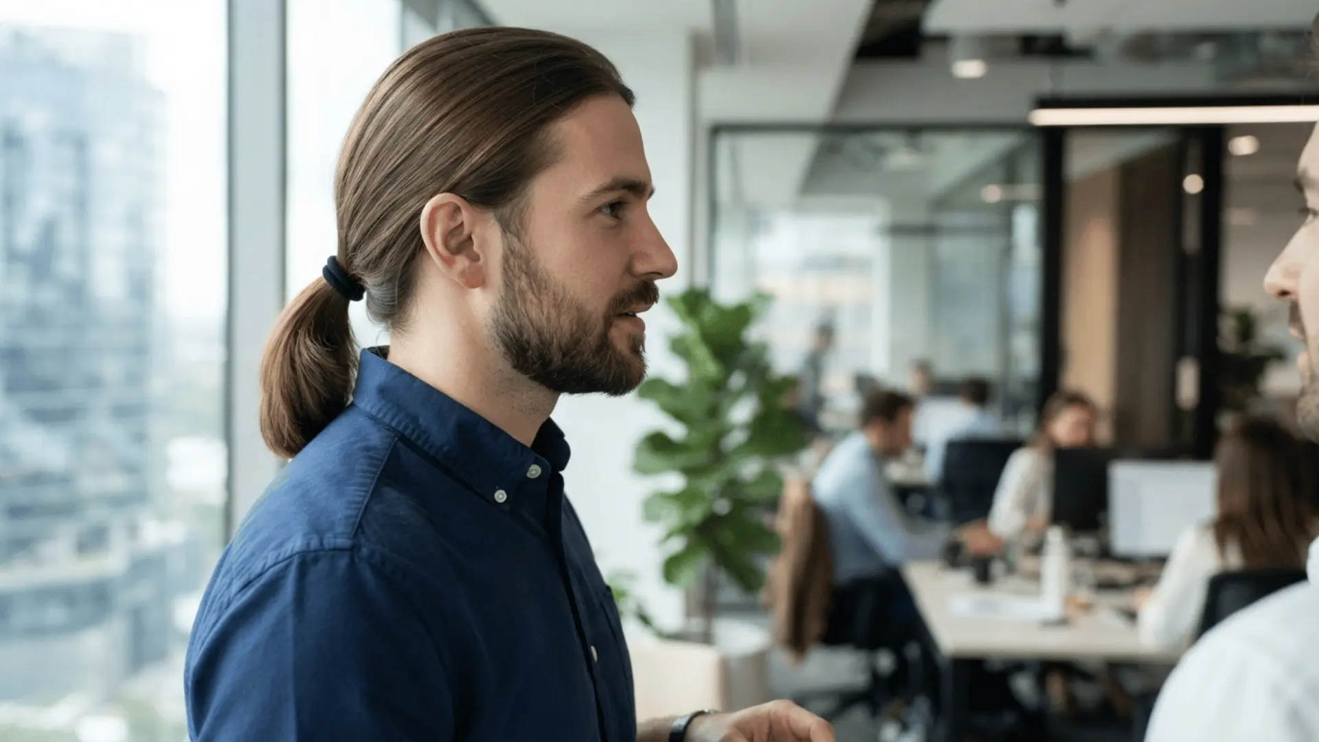 man with a ponytail talks to another person in a bright modern office while coworkers work in the background