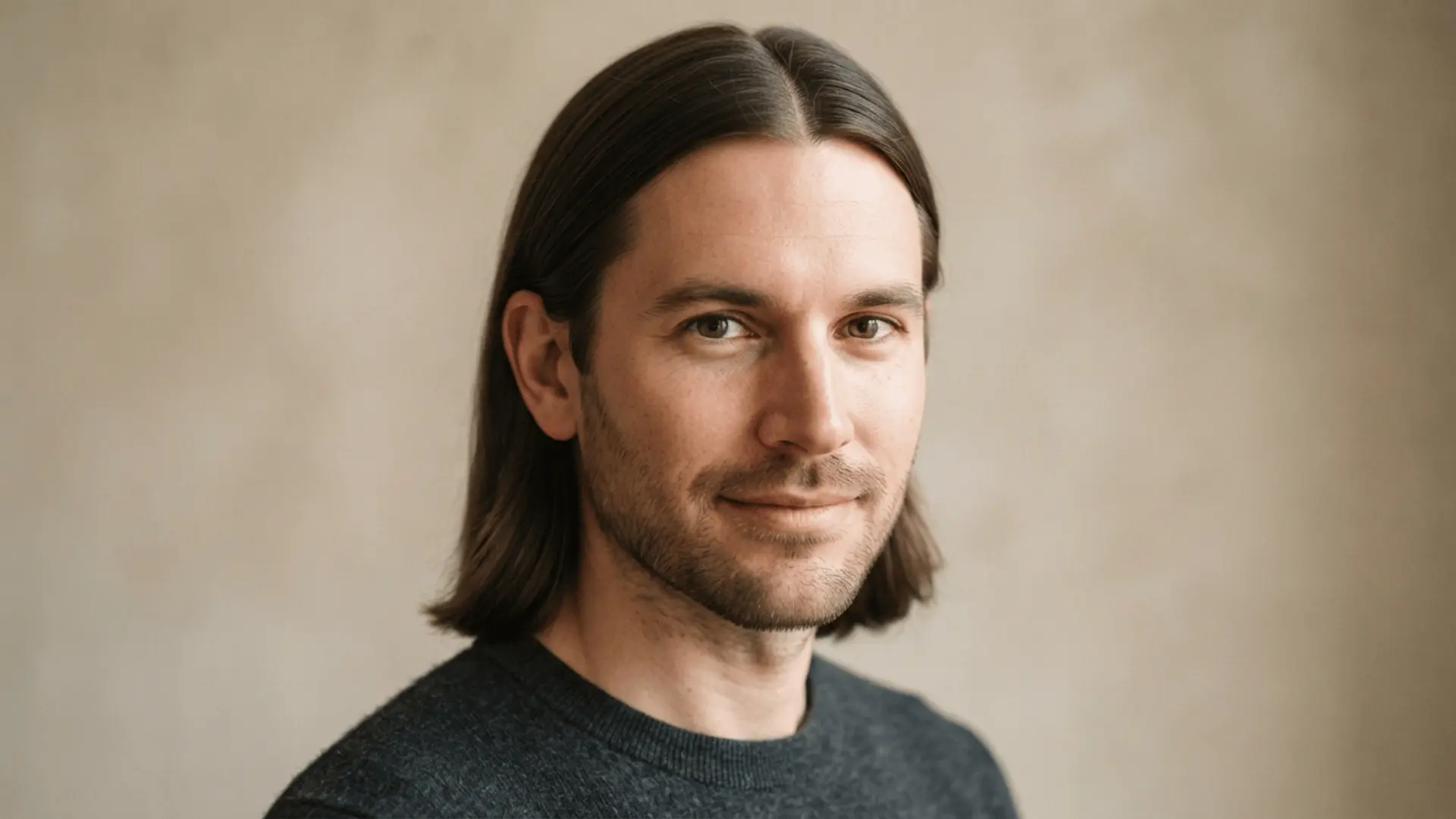 close portrait of a man with shoulder length brown hair and a slight smile against a plain beige background