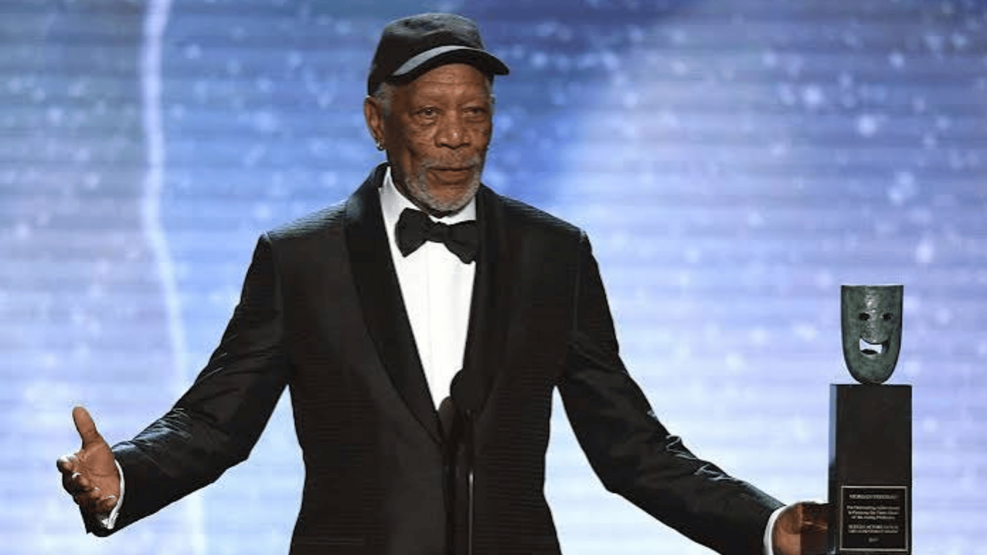 Morgan Freeman wearing a tuxedo and cap while holding an award on stage at an event.
