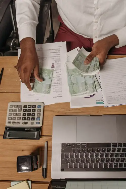 Person counting euro bills at wooden desk with laptop and calculator