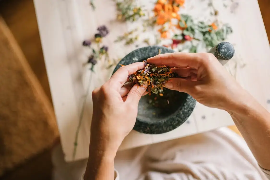 Hands mixing dried flowers with a pestle on a wooden table indoors