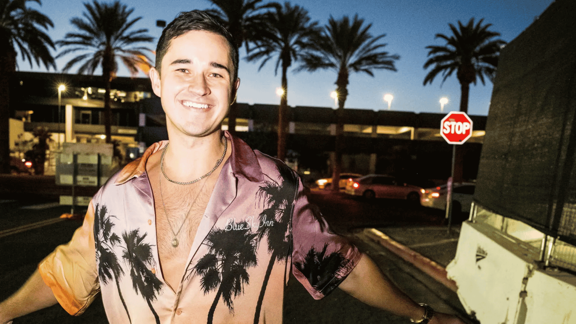 Smiling DJ in palm-print shirt outside a city venue at dusk