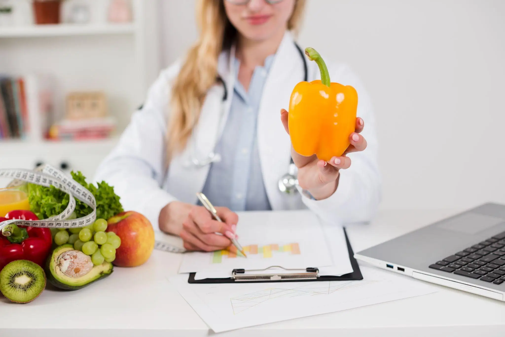 Doctor in white coat holding yellow bell pepper at desk with fresh fruits and laptop