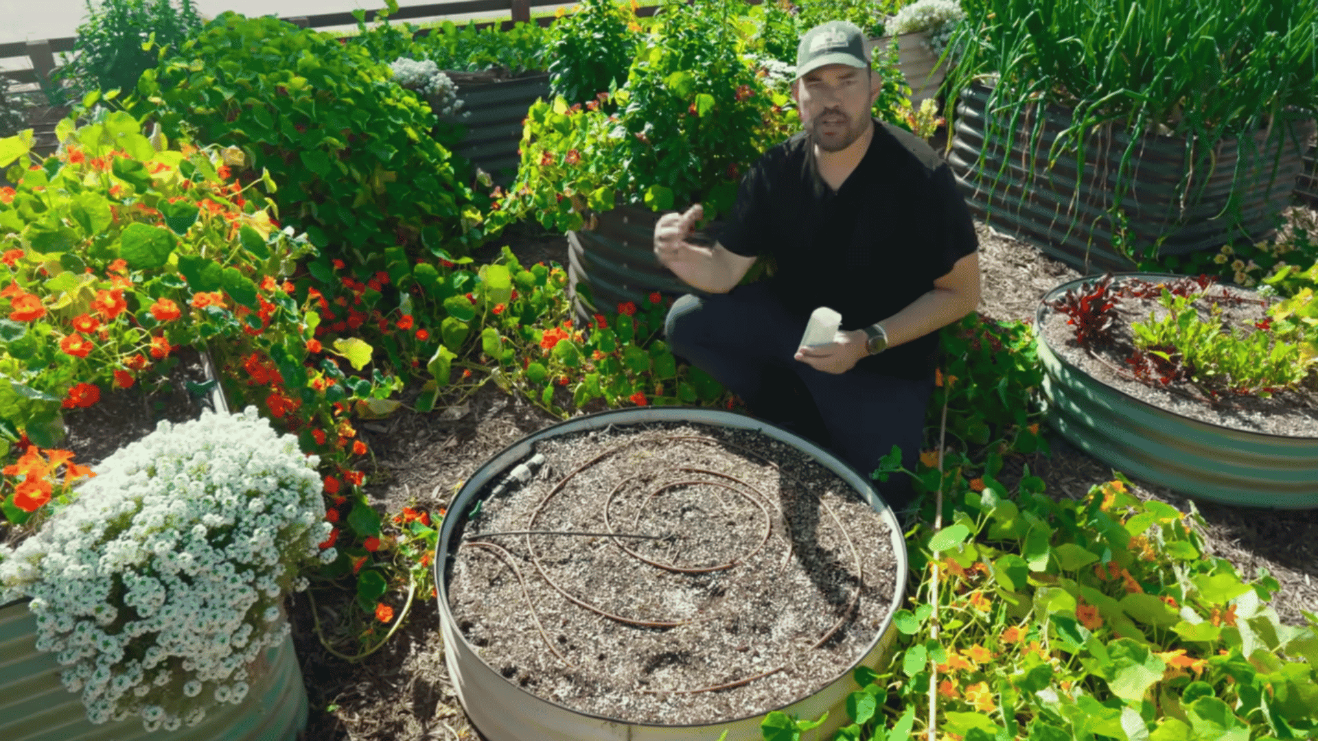 man kneels beside a round raised garden bed holding a small container while explaining coiled irrigation tubing among flowers