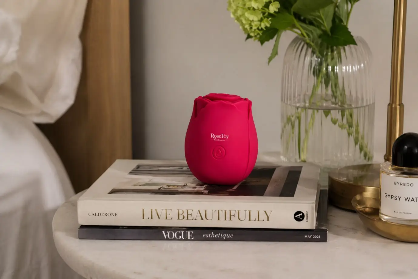 Red rose-shaped object on stacked books next to perfume bottle and flower vase on bedside table