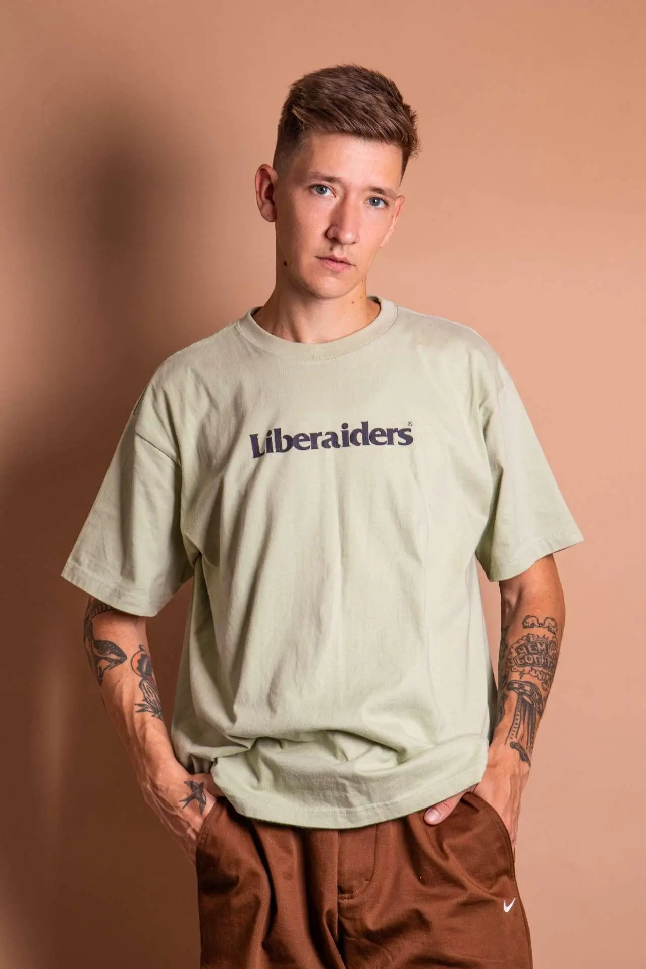 Young person in beige t-shirt with tattoos, standing against a light brown background