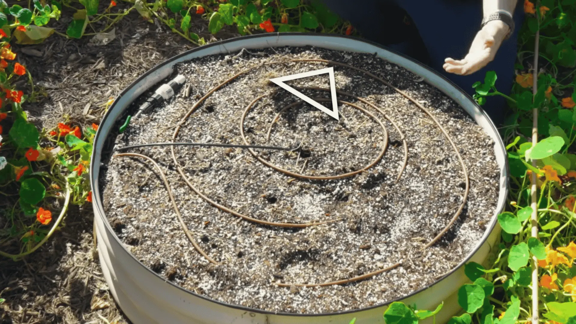 round raised garden bed holds coiled irrigation tubing with a white triangular marker resting on the dry soil