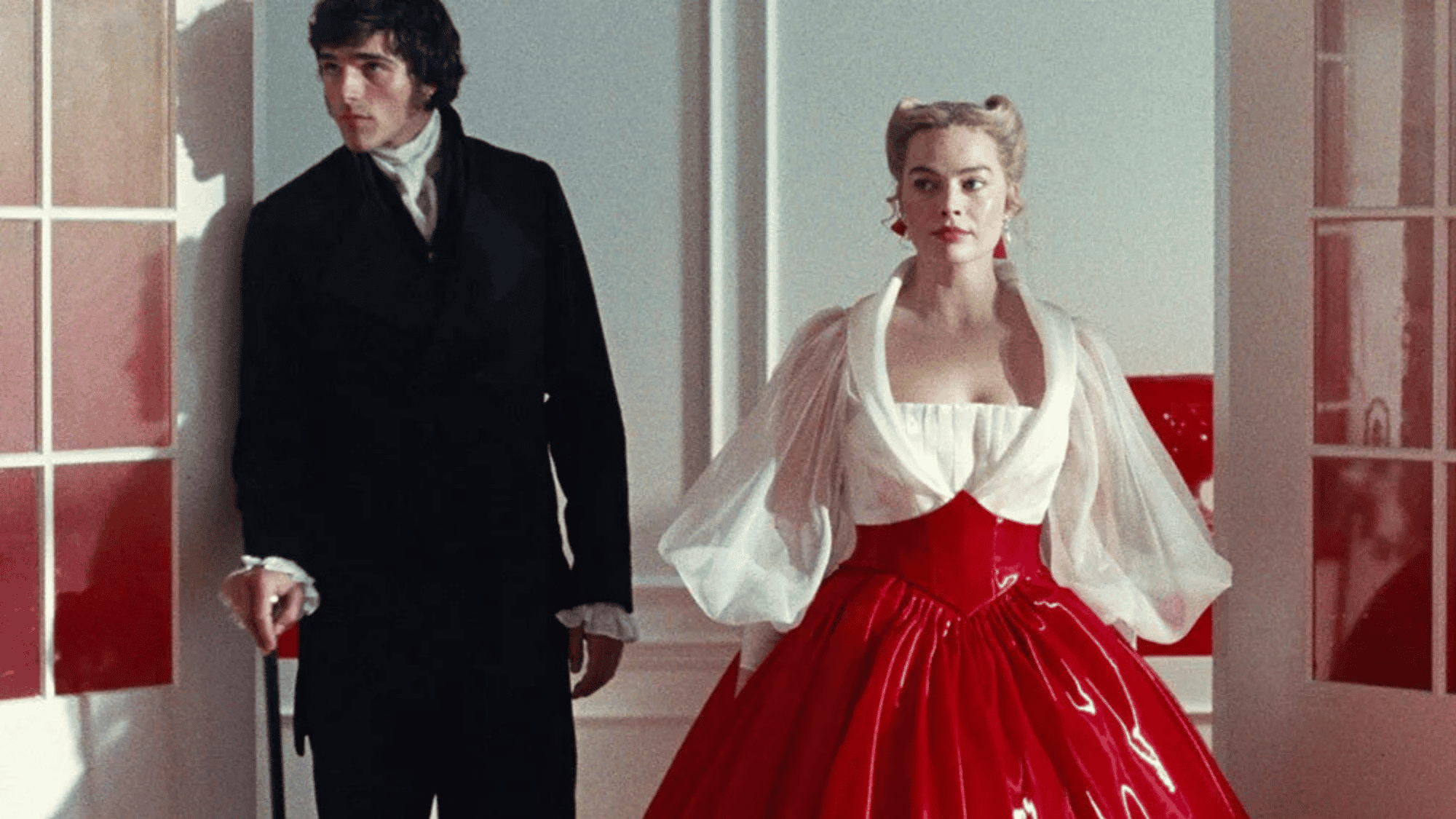 Period drama pair stands in a bright room as woman wears a bold red gown