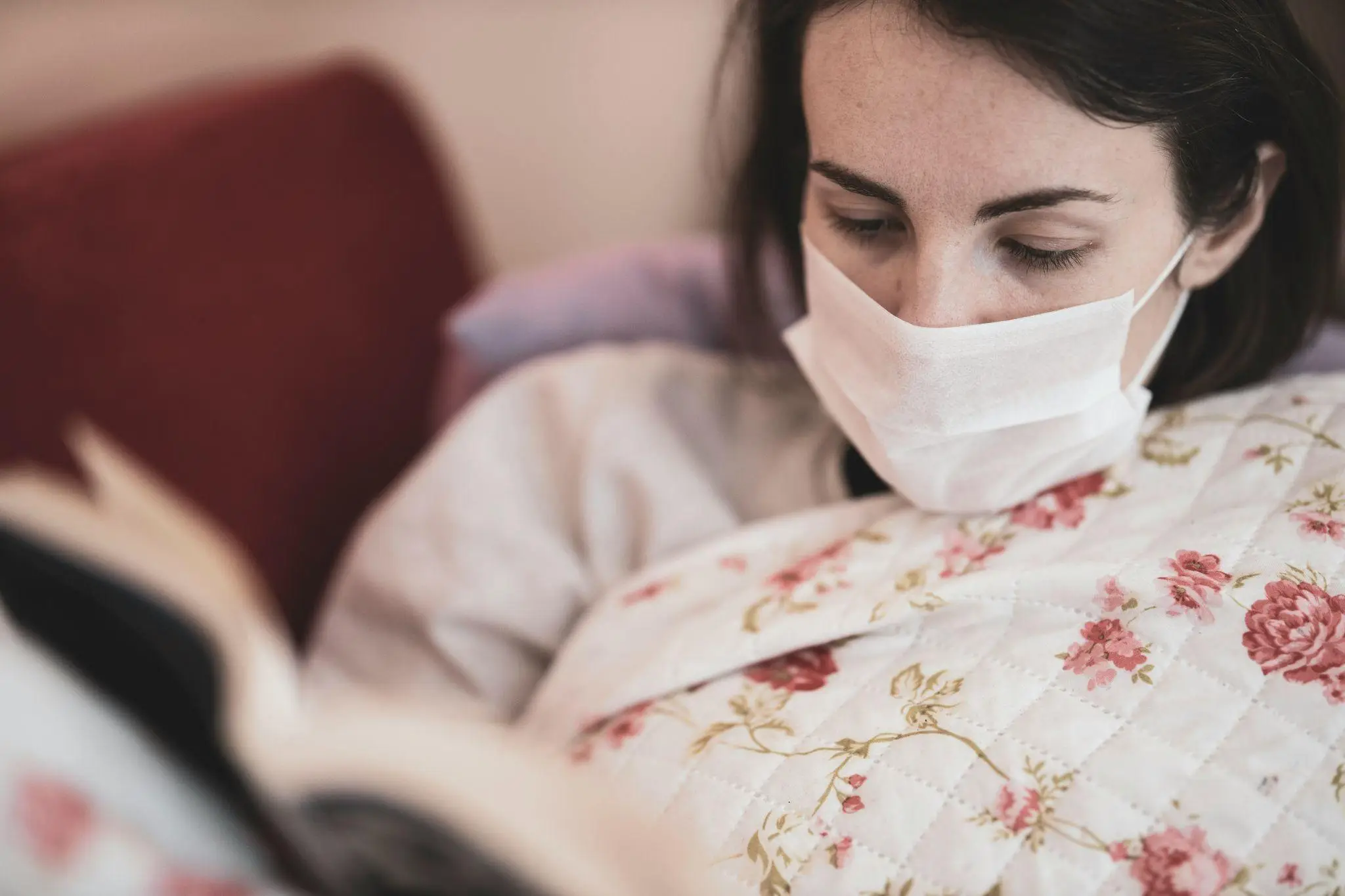 Person wearing a mask reading a book under a floral quilt indoors