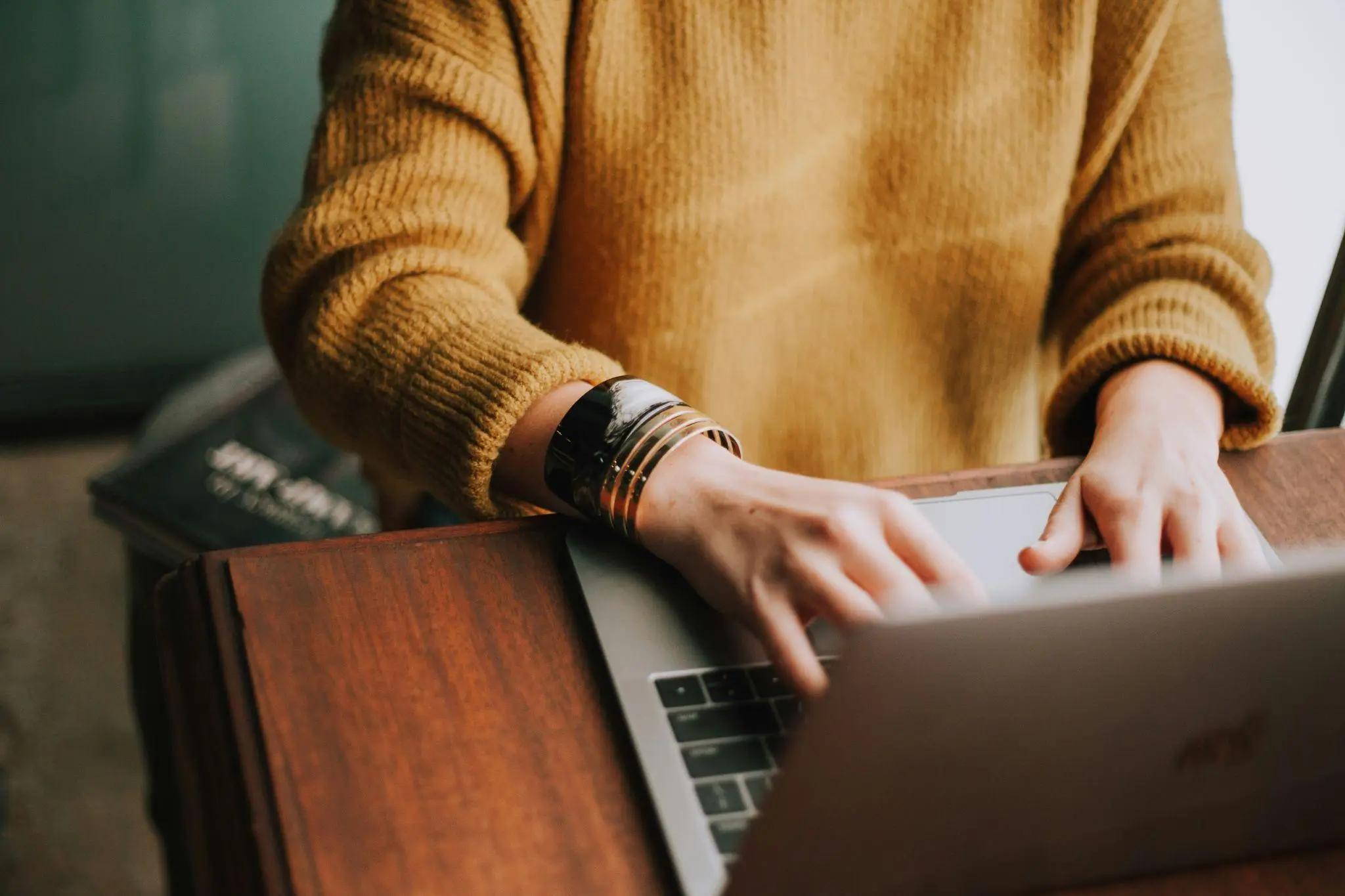 Person typing on a laptop wearing a mustard sweater at a wooden desk