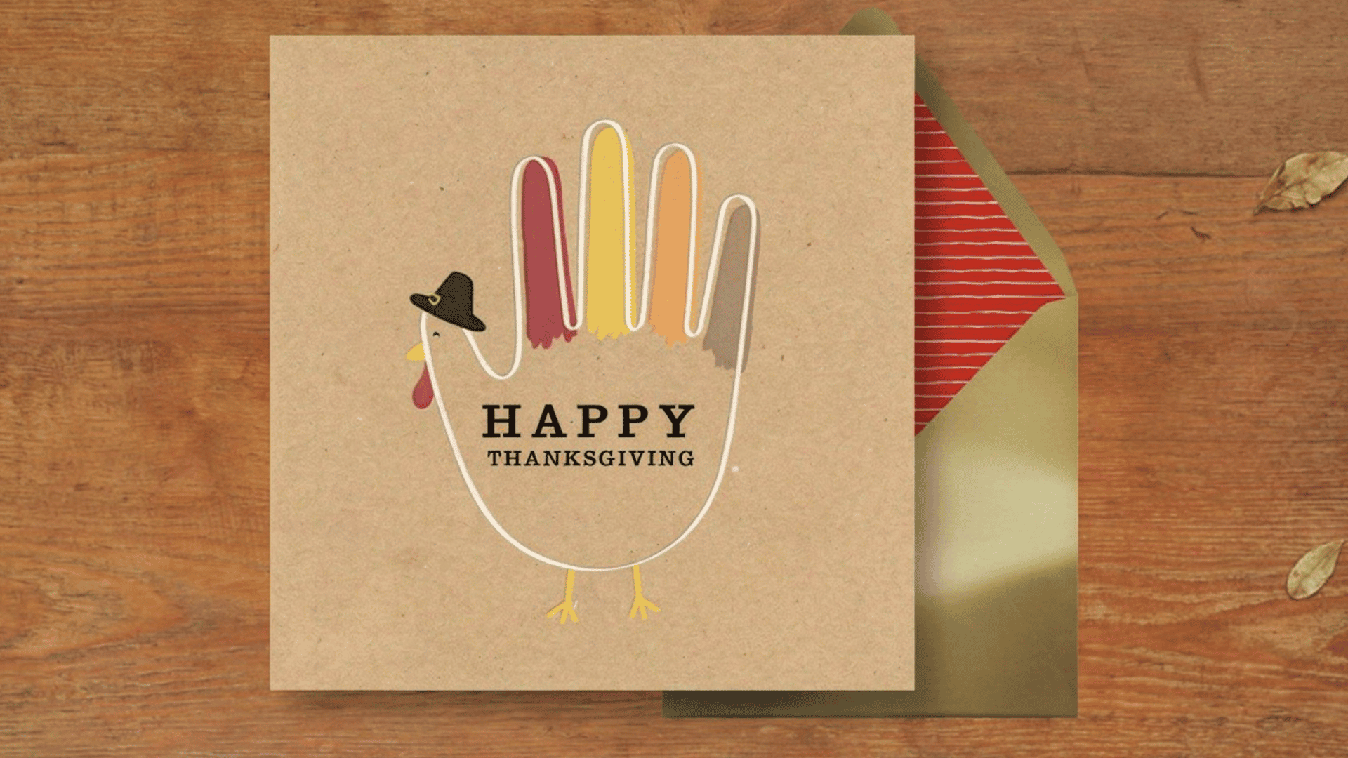 thanksgiving card with a hand shaped turkey beside a green envelope on a wooden table with leaves and twine
