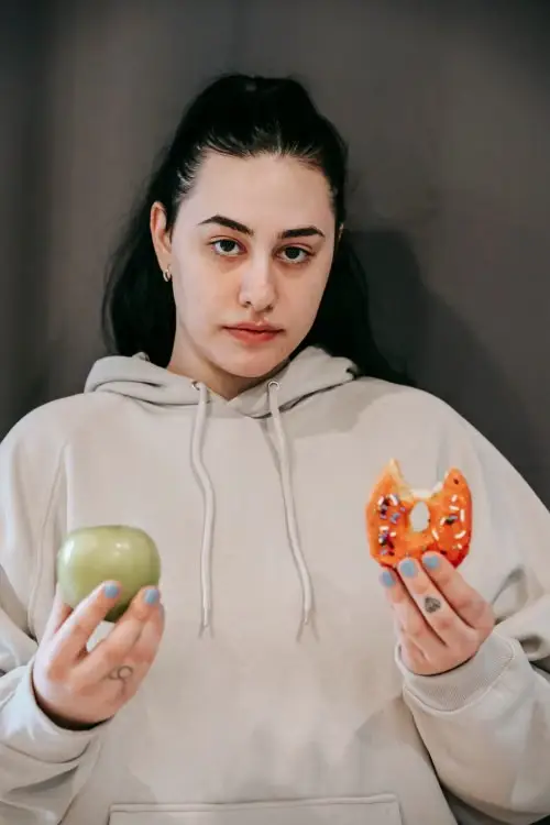 Person in beige hoodie holding a green apple and a bitten donut indoors