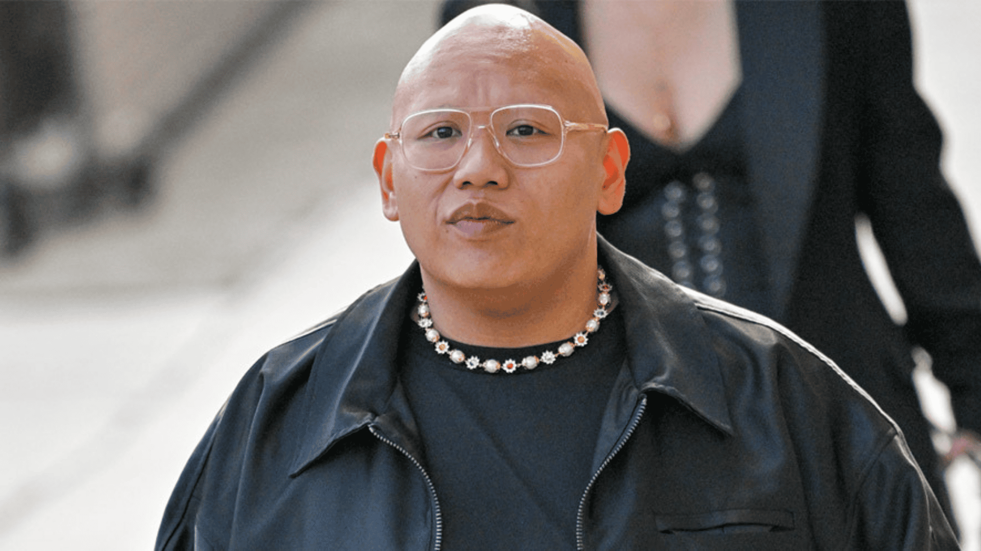 Bald actor in clear glasses wearing a black jacket and floral necklace outdoors