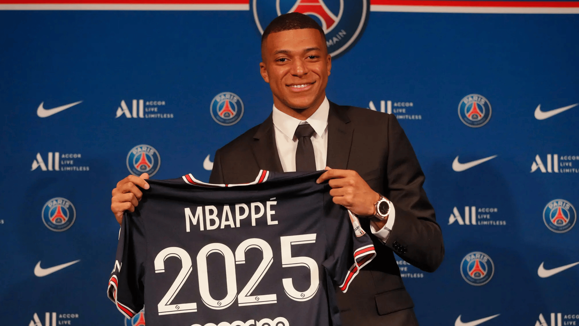 Kylian Mbappé smiles while holding a PSG jersey during his unveiling at an official club presentation.