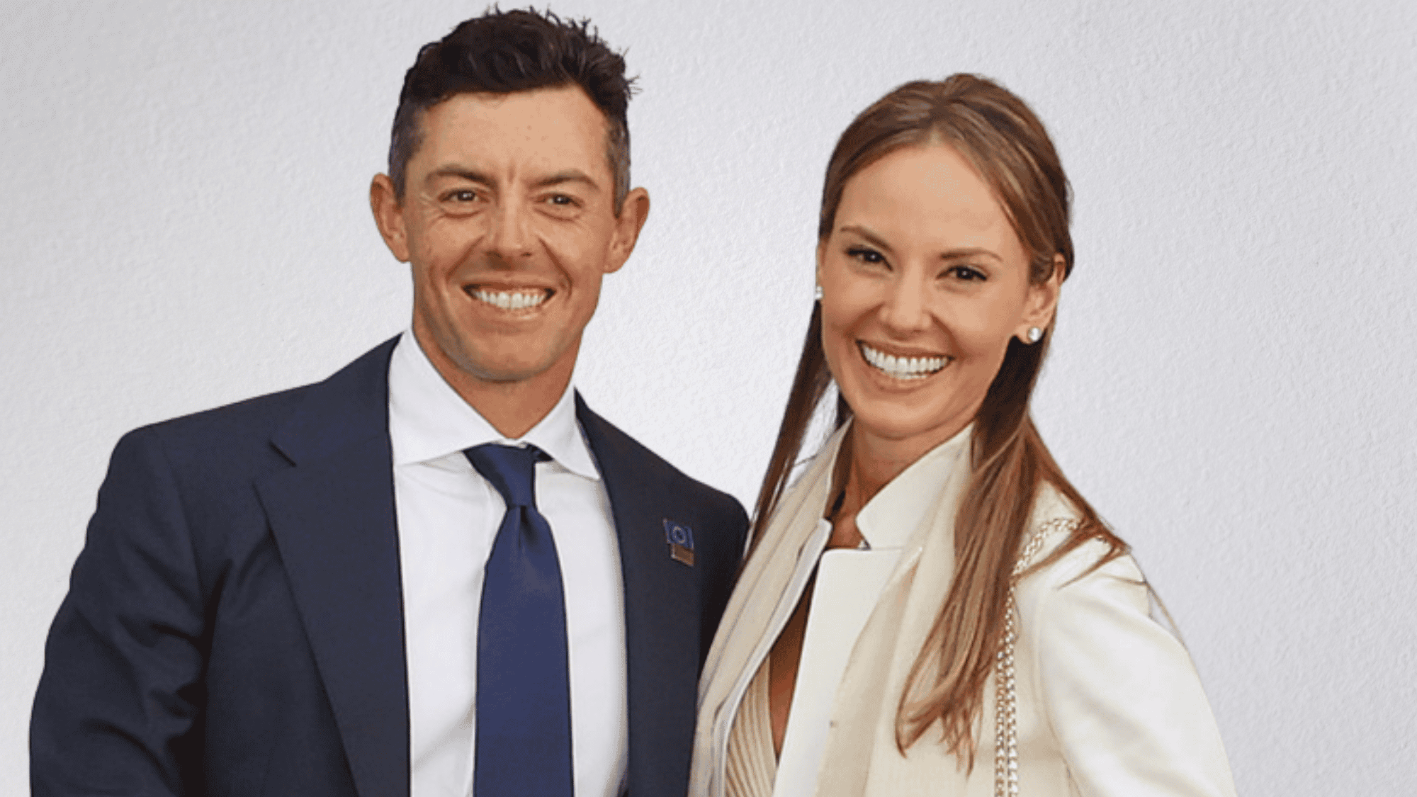 Rory McIlroy in a dark suit and blue tie smiling next to Erica Stoll in a white blazer.