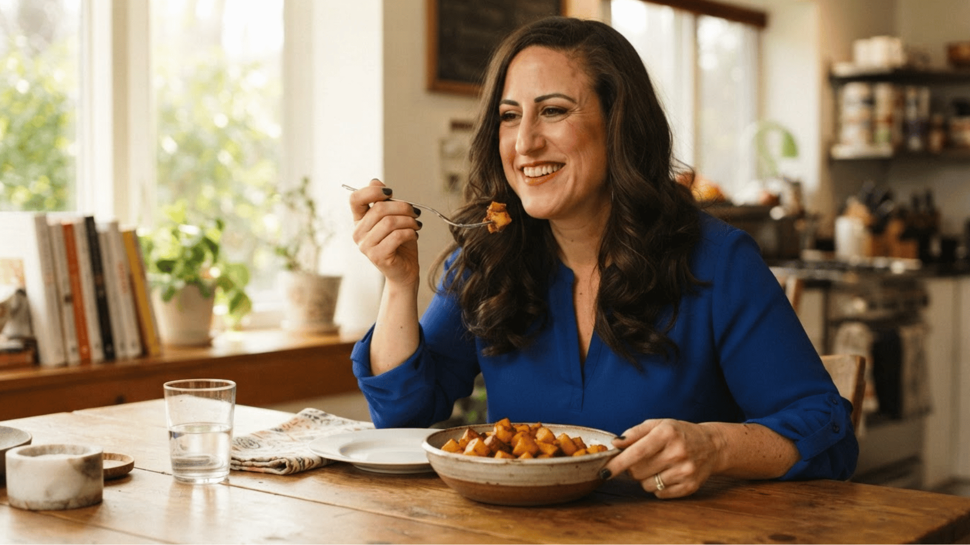 woman smiling while eating roasted butternut squash at a wooden dining table in a bright home kitchen