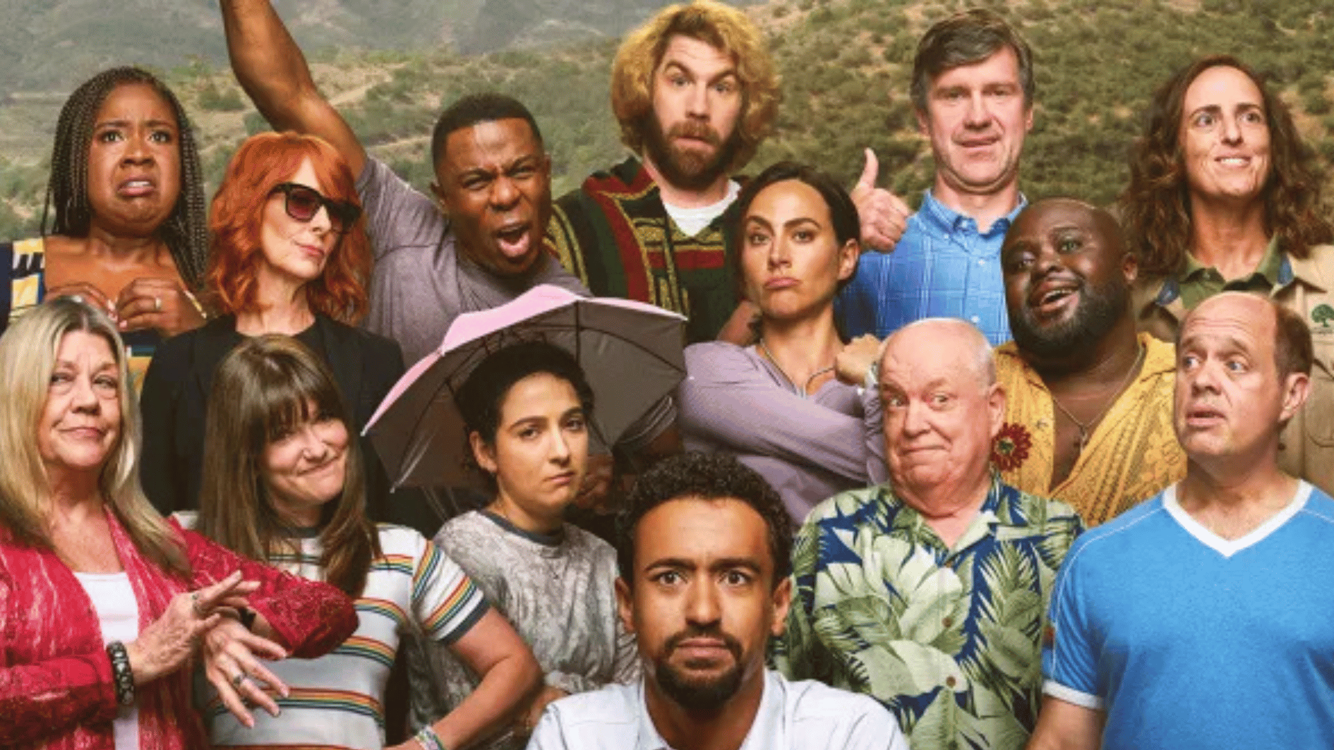 a large comedy cast poses together, making playful expressions against a mountain backdrop