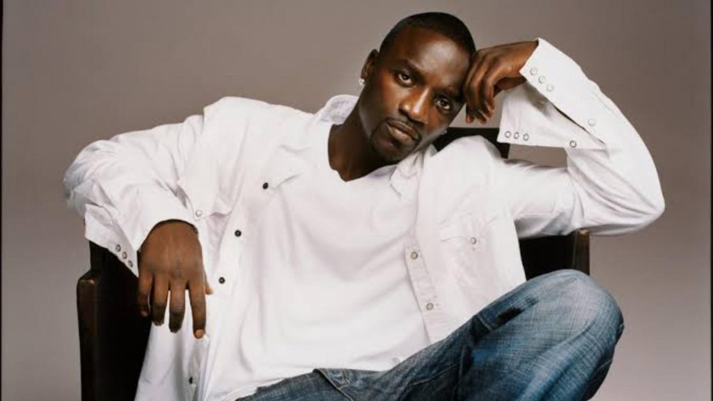 Singer Akon leaning back in a wooden chair wearing a white unbuttoned shirt over a white t-shirt and blue jeans.