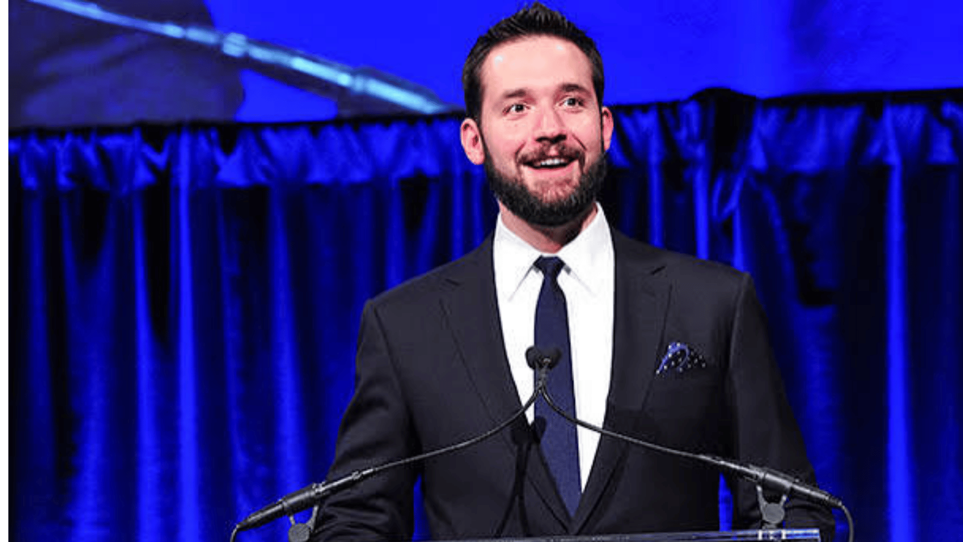 Alexis Ohanian speaking at an event while wearing a suit.