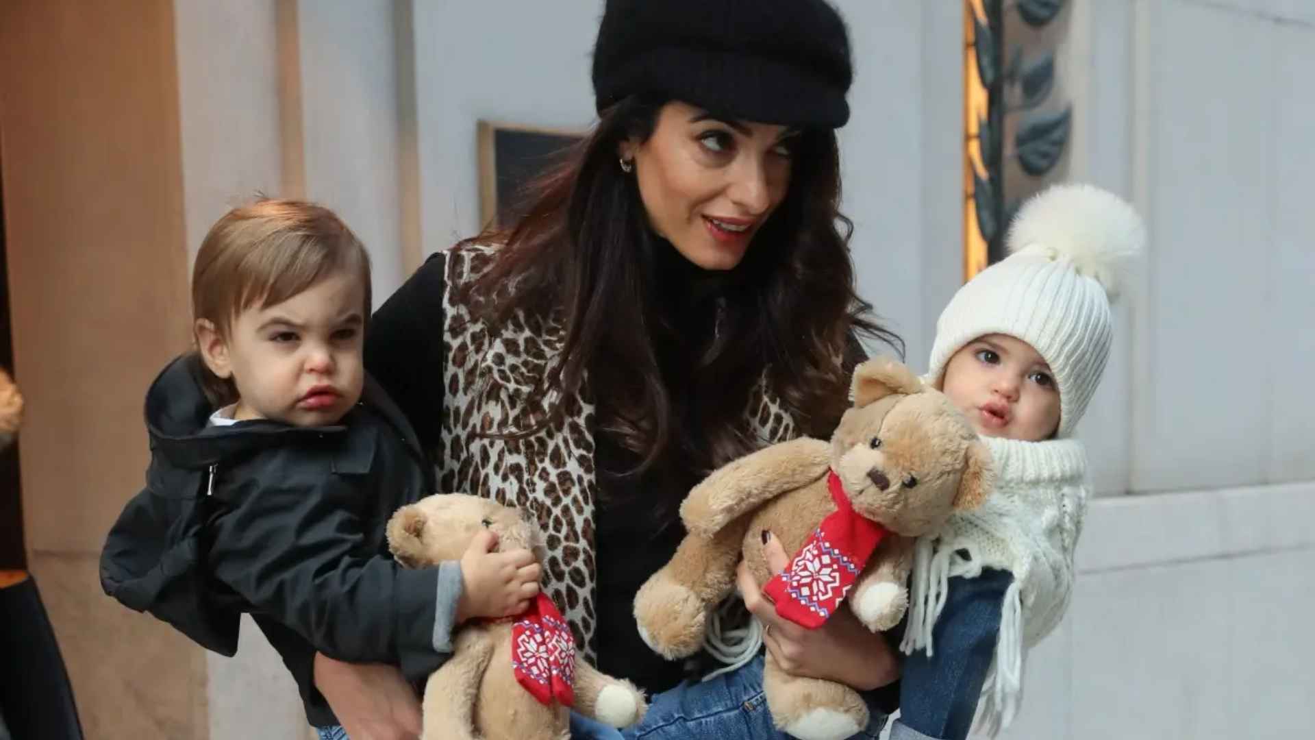 Amal Clooney holds her twins wearing winter clothing while they carry teddy bears indoors during a family moment