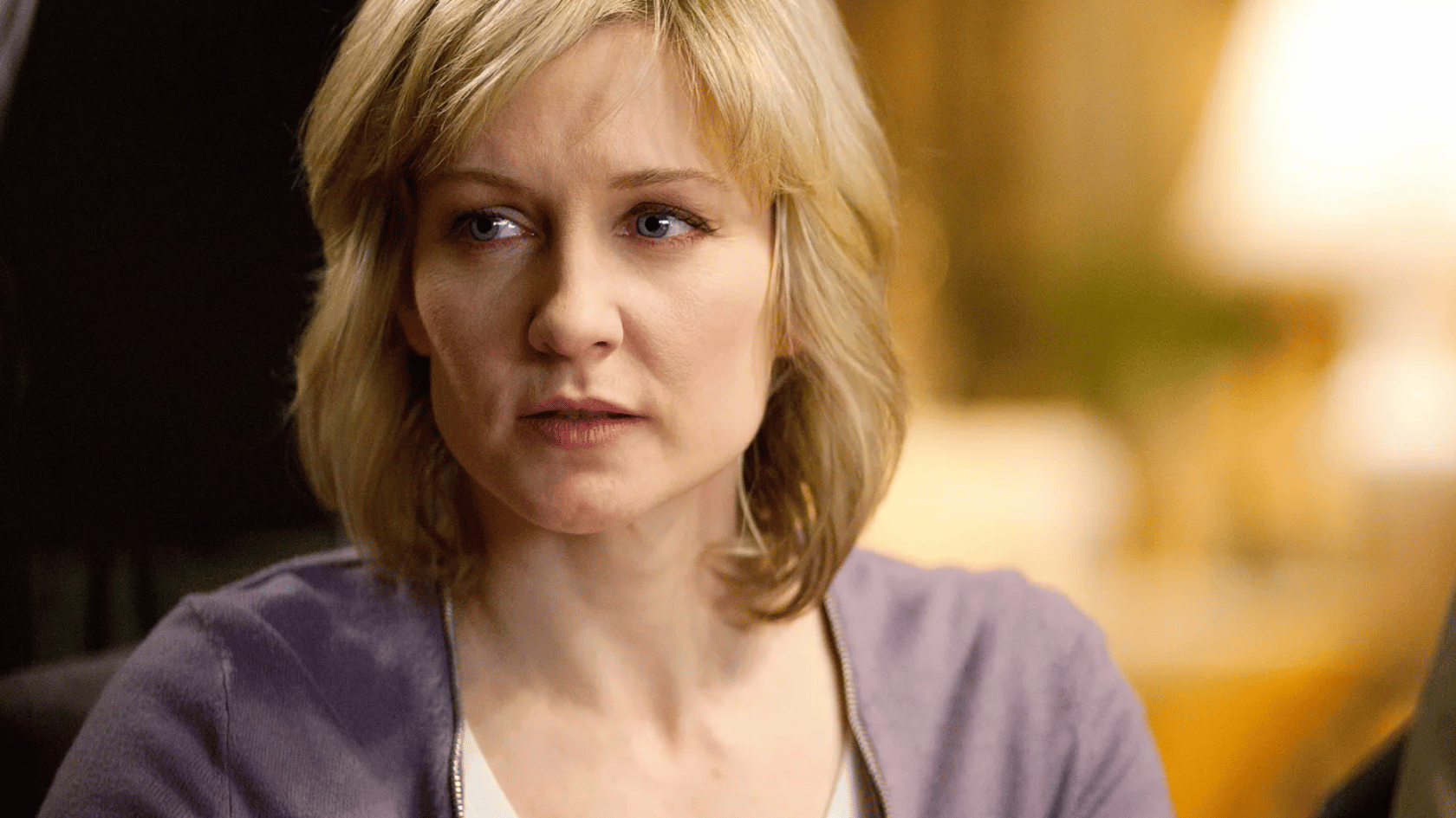 amy carlson with blonde hair sits indoors wearing a purple cardigan, looking to the side with a serious expression in warm lighting