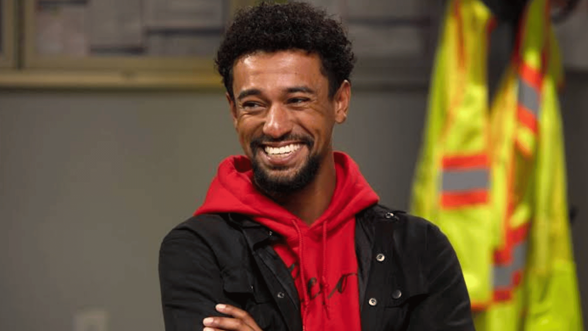 anthony wearing a red hoodie and black jacket stands indoors with safety vests visible in the background