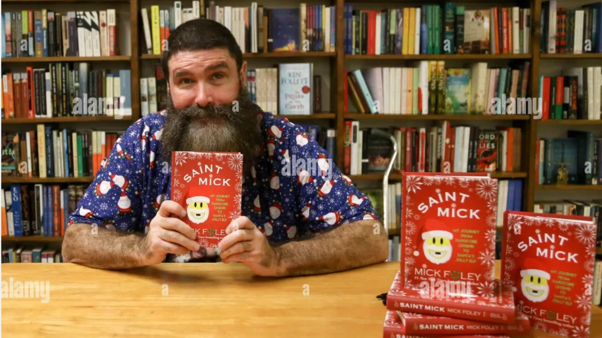 bearded man holding a saint mick book at a bookstore table with copies displayed beside him and bookshelves behind him