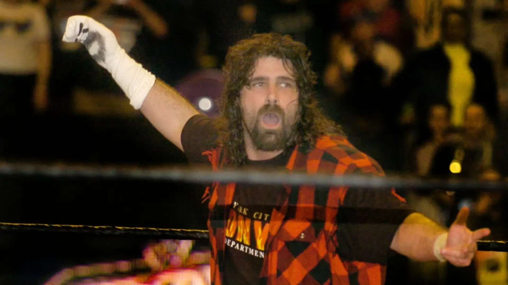 bearded wrestler in red and black plaid vest raises one arm in the wrestling ring as a blurred crowd watches behind him