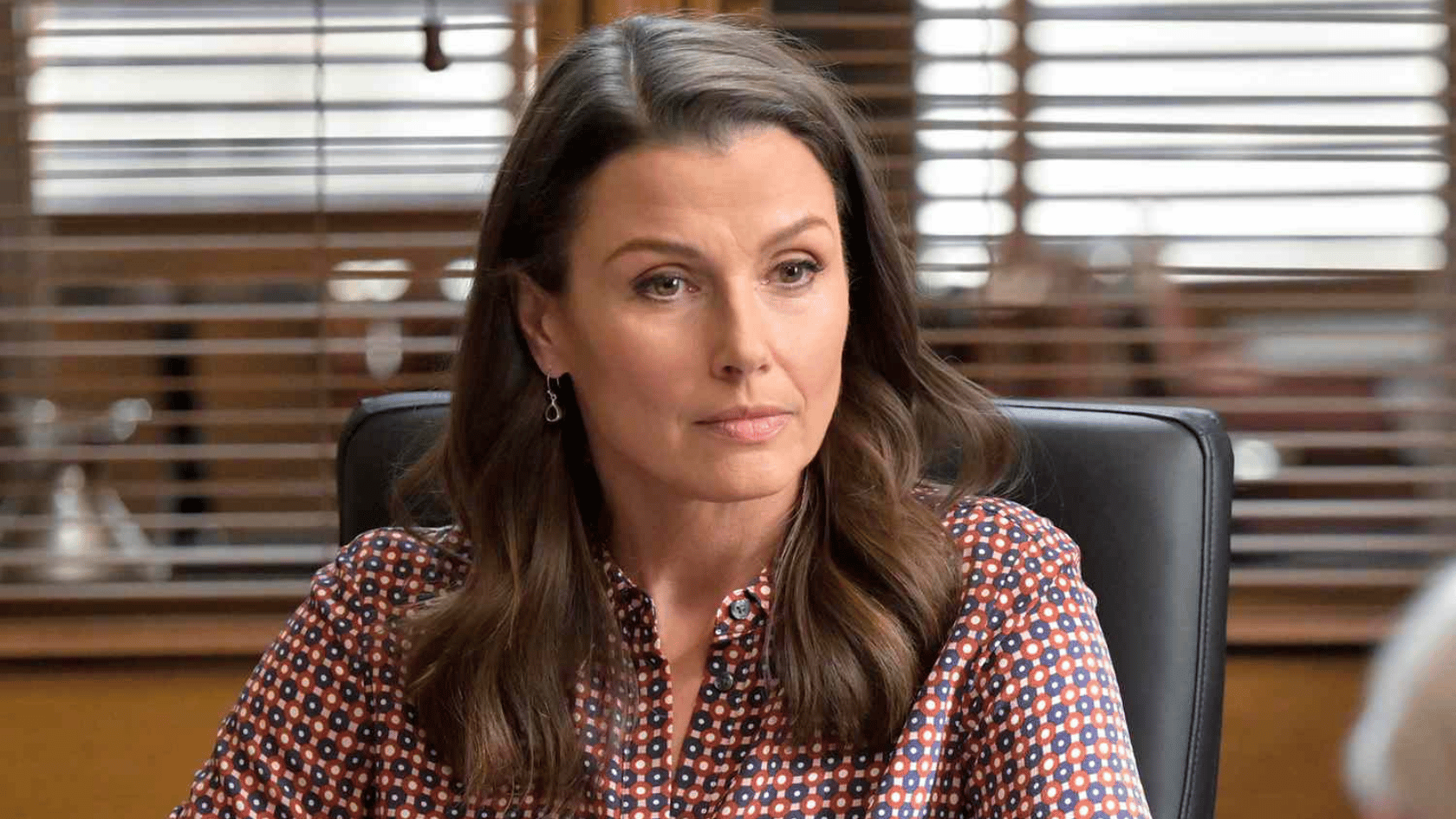 bridget moynahan sitting in an office chair, wearing a patterned blouse and looking to the sid