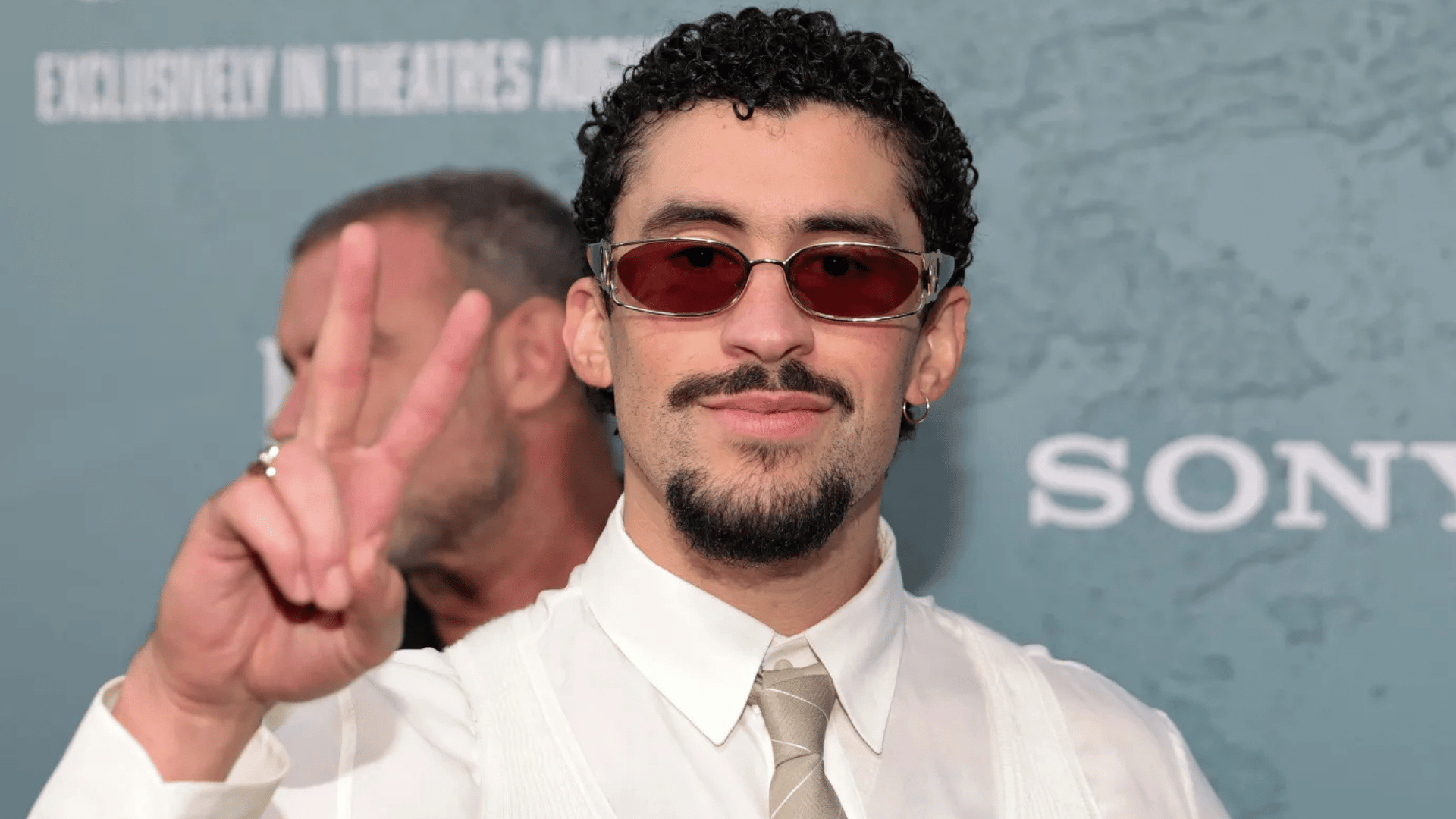 Bad Bunny wearing red sunglasses and making a peace sign at a public event in LA