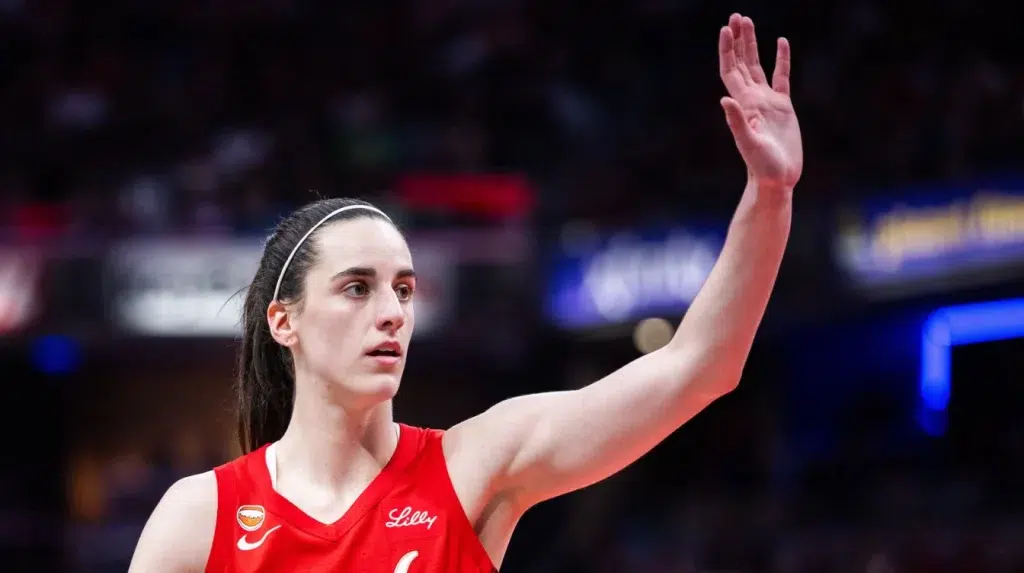 caitlin clark in a red basketball jersey raising her hand on court with a blurred arena background