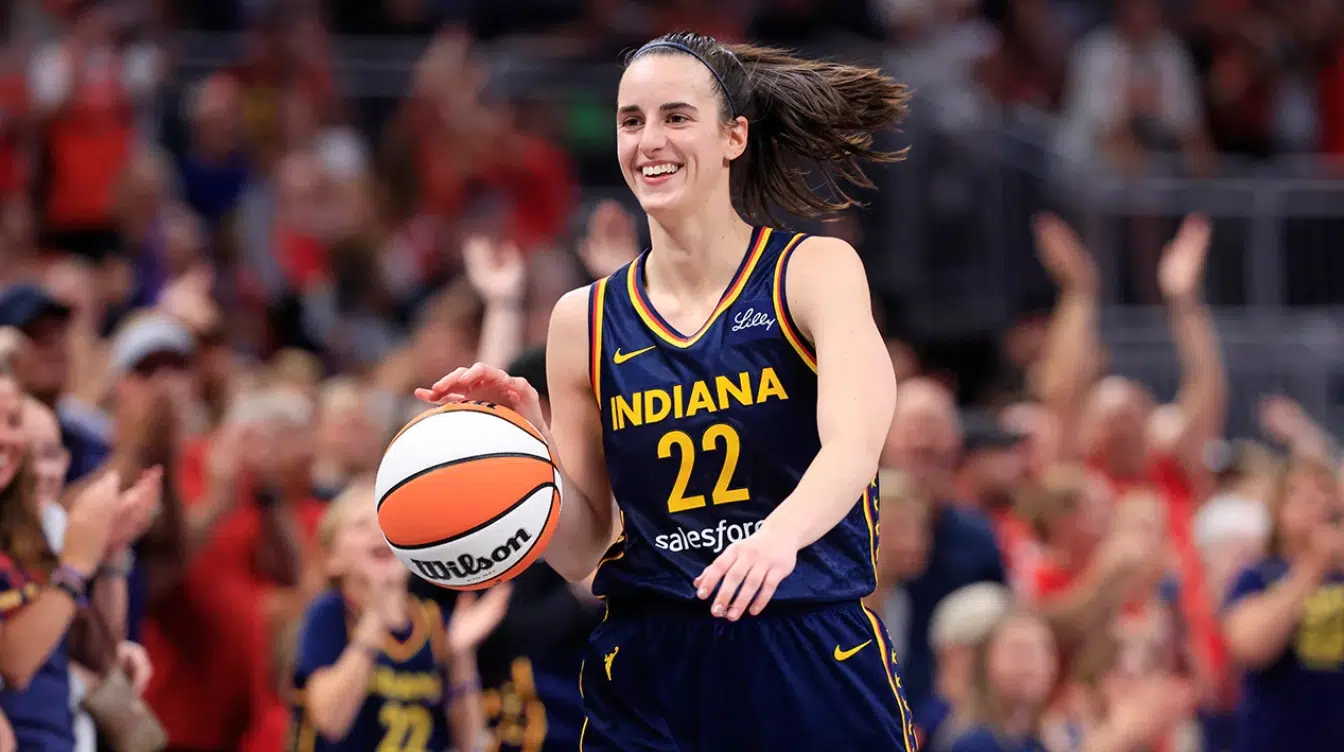 caitlin clark smiling in an indiana fever jersey while holding a basketball on court with cheering fans behind her
