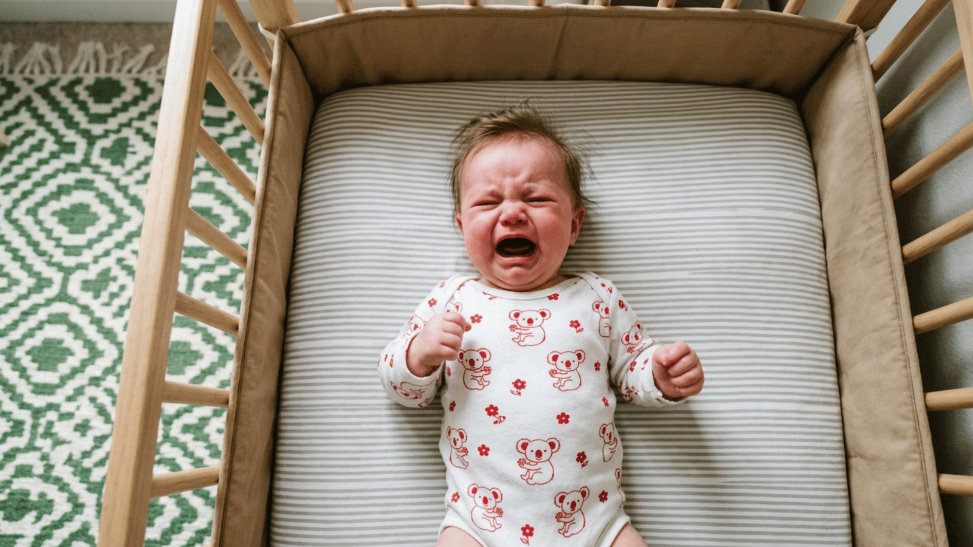 an upset, crying baby wearing a koala-print onesie lies on its back in a wooden crib with striped bedding