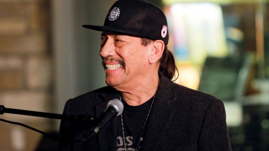 Danny Trejo smiling in profile at a microphone, wearing a black Trejo's Coffee & Donuts cap and dark blazer