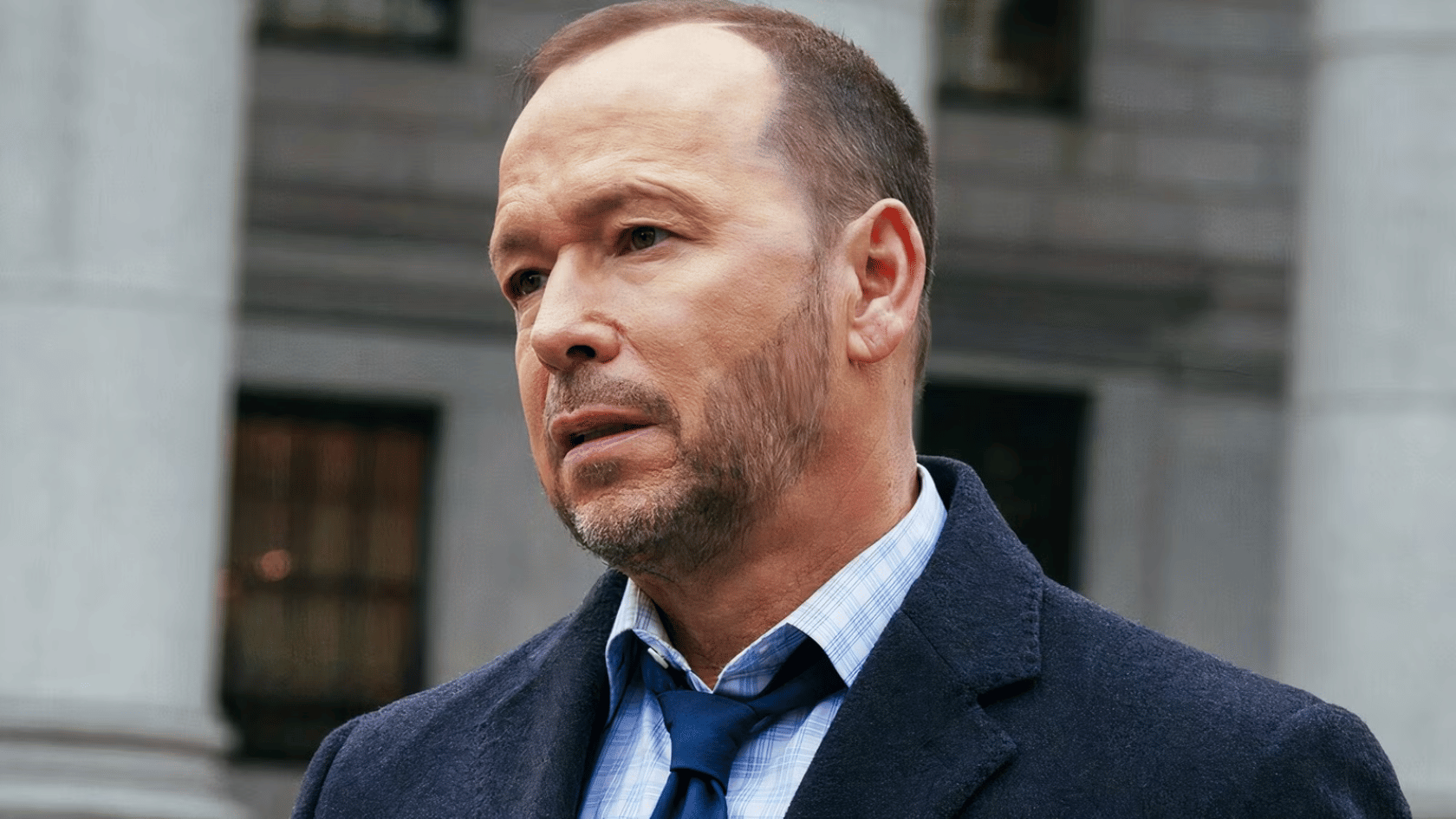 donnie wahlberg wearing a navy coat and blue tie stands outdoors in front of a building, looking to the side with a serious expression
