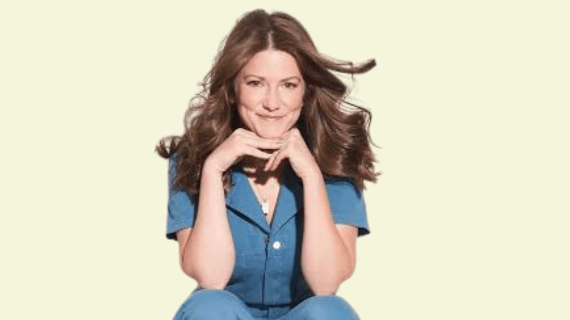 emily with long brown hair wearing a blue outfit resting her chin on her hands against a light background