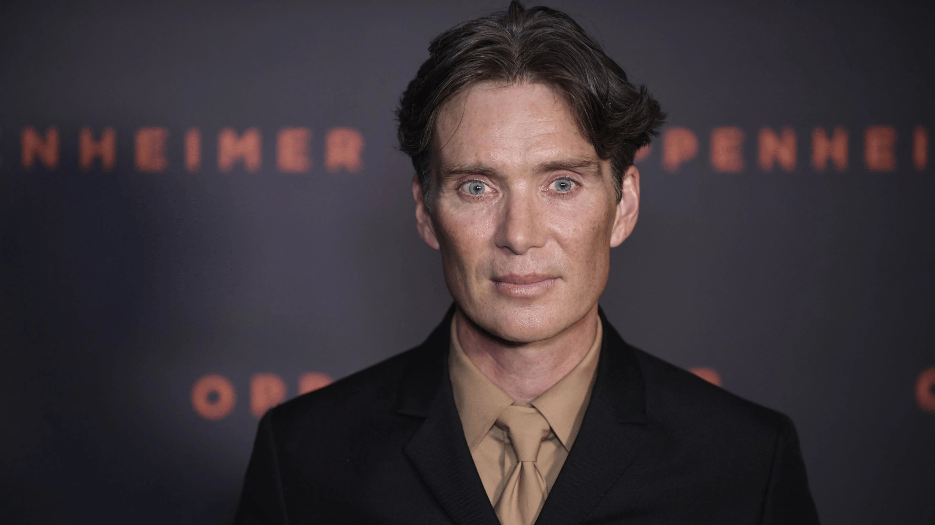cillian murphy in a black suit and tan shirt at an oppenheimer premiere backdrop