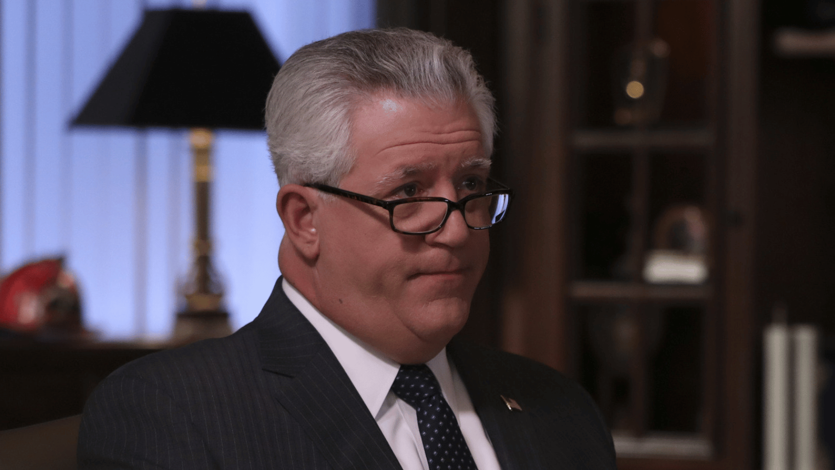 gregory jbara with glasses wearing a dark suit and tie sits indoors, looking to the side with a serious expression in a formal office setting