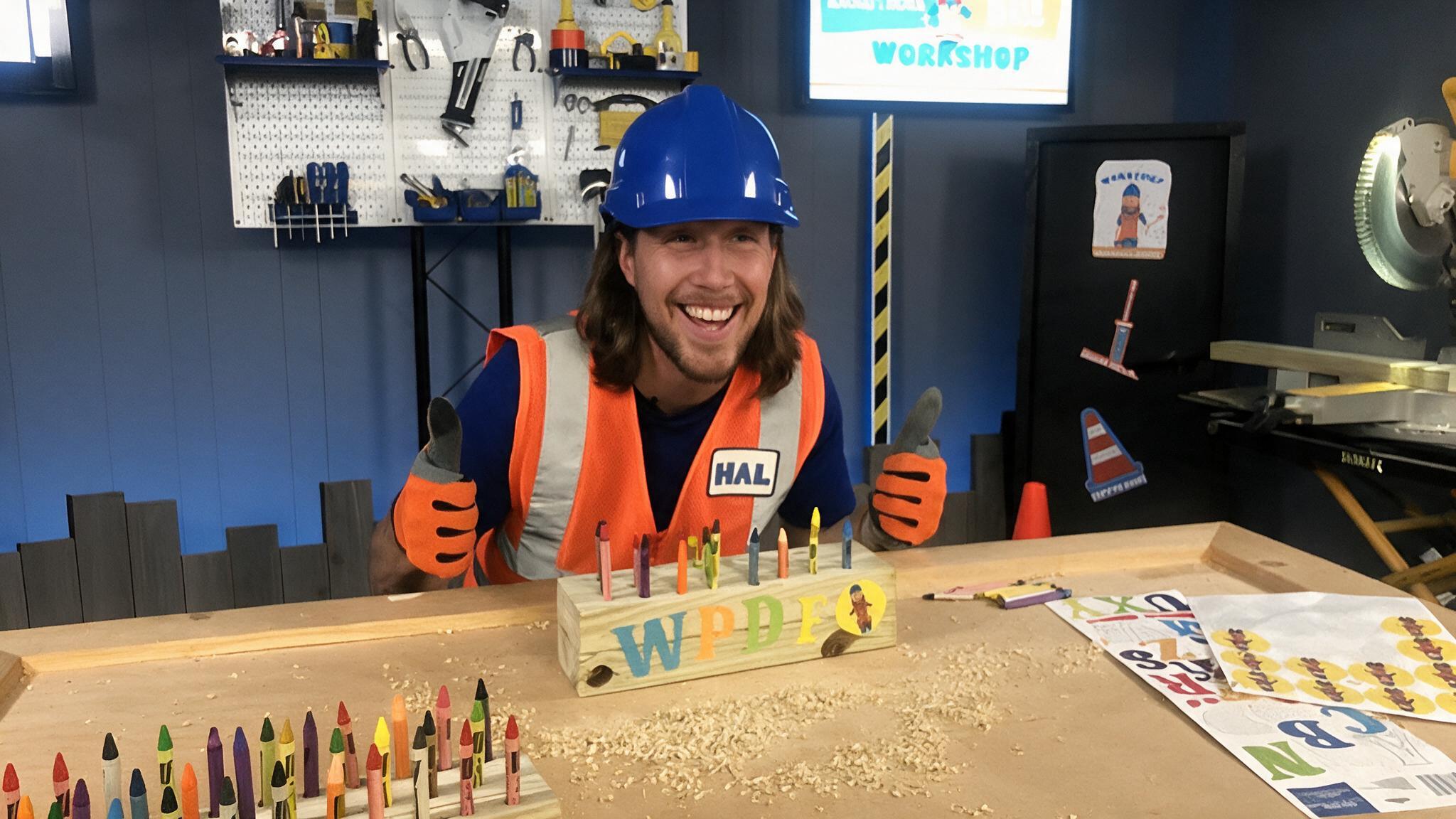 handyman hal wearing a blue helmet and orange safety vest smiling in a workshop with tools, crayons, and woodworking materials on a table
