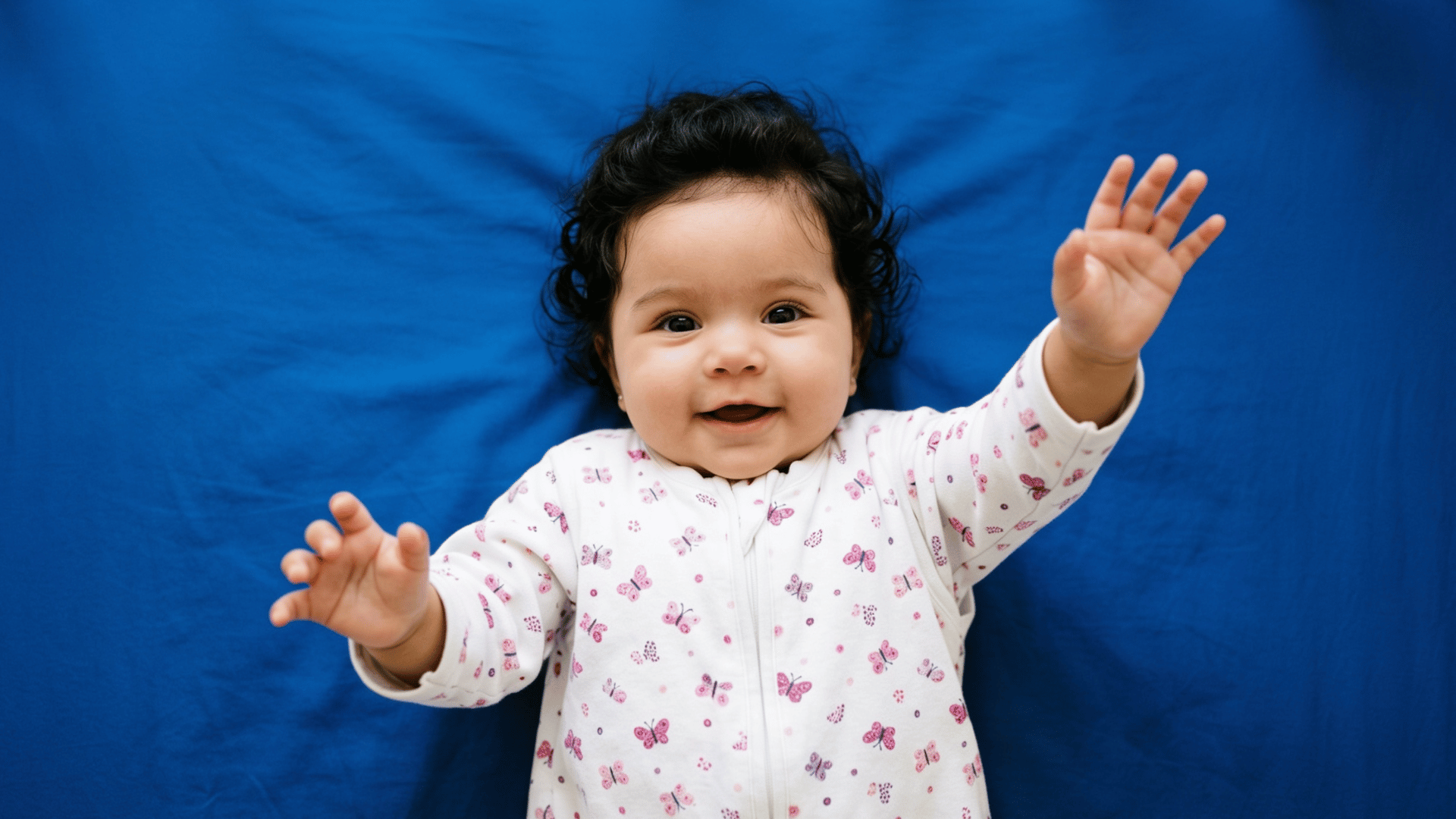 a happy, smiling baby with dark curly hair lies on a blue blanket, reaching up with both of their hands