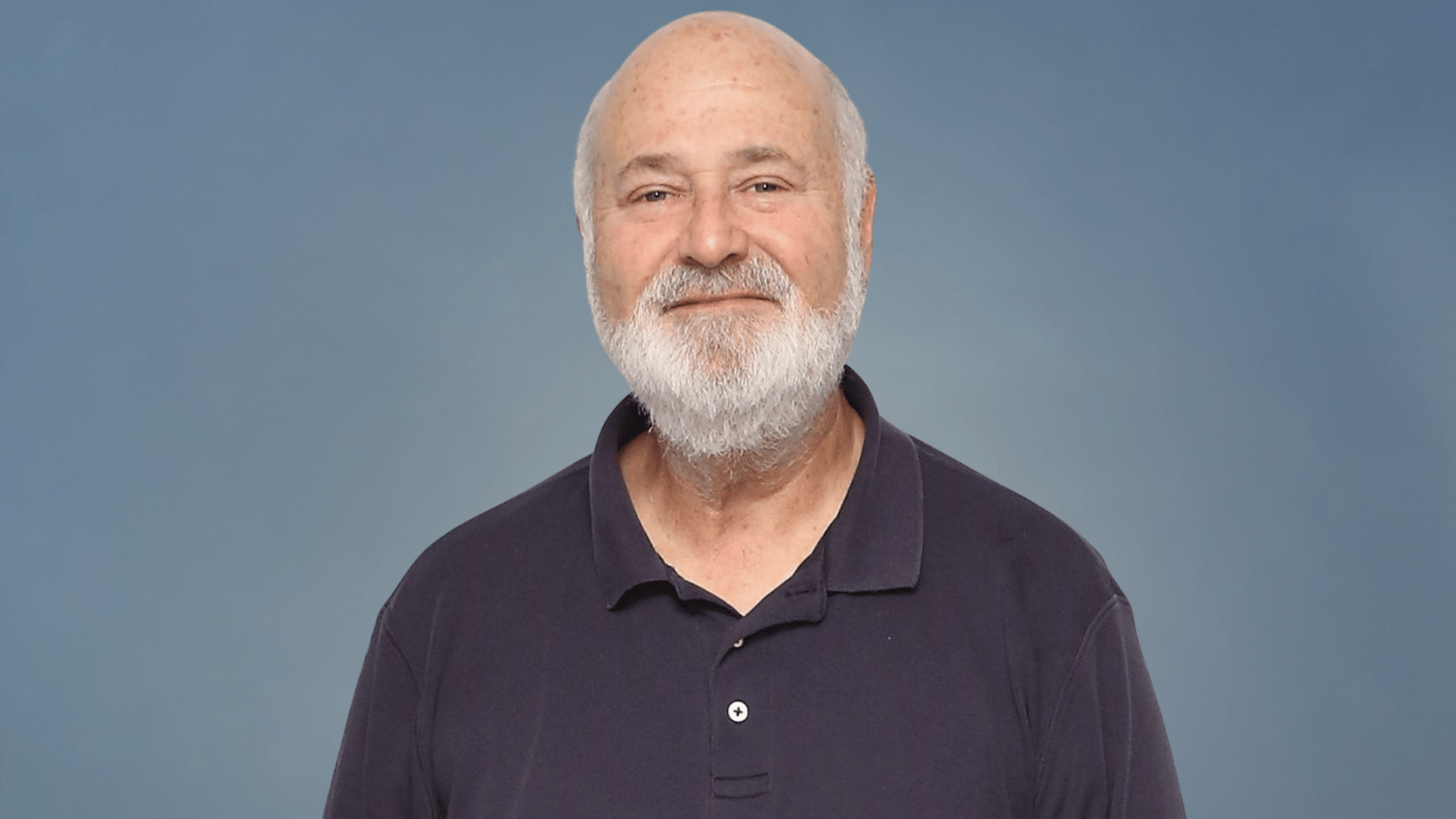 Rob Reiner smiling in a black polo shirt against a blue background