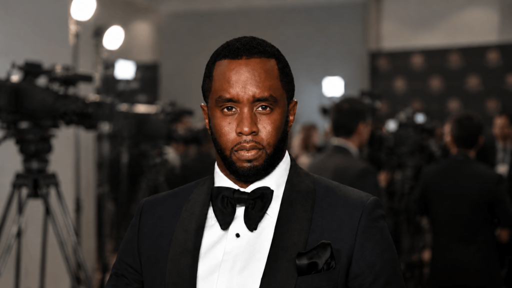 Sean Combs in a tuxedo at a media interview event with cameras behind him
