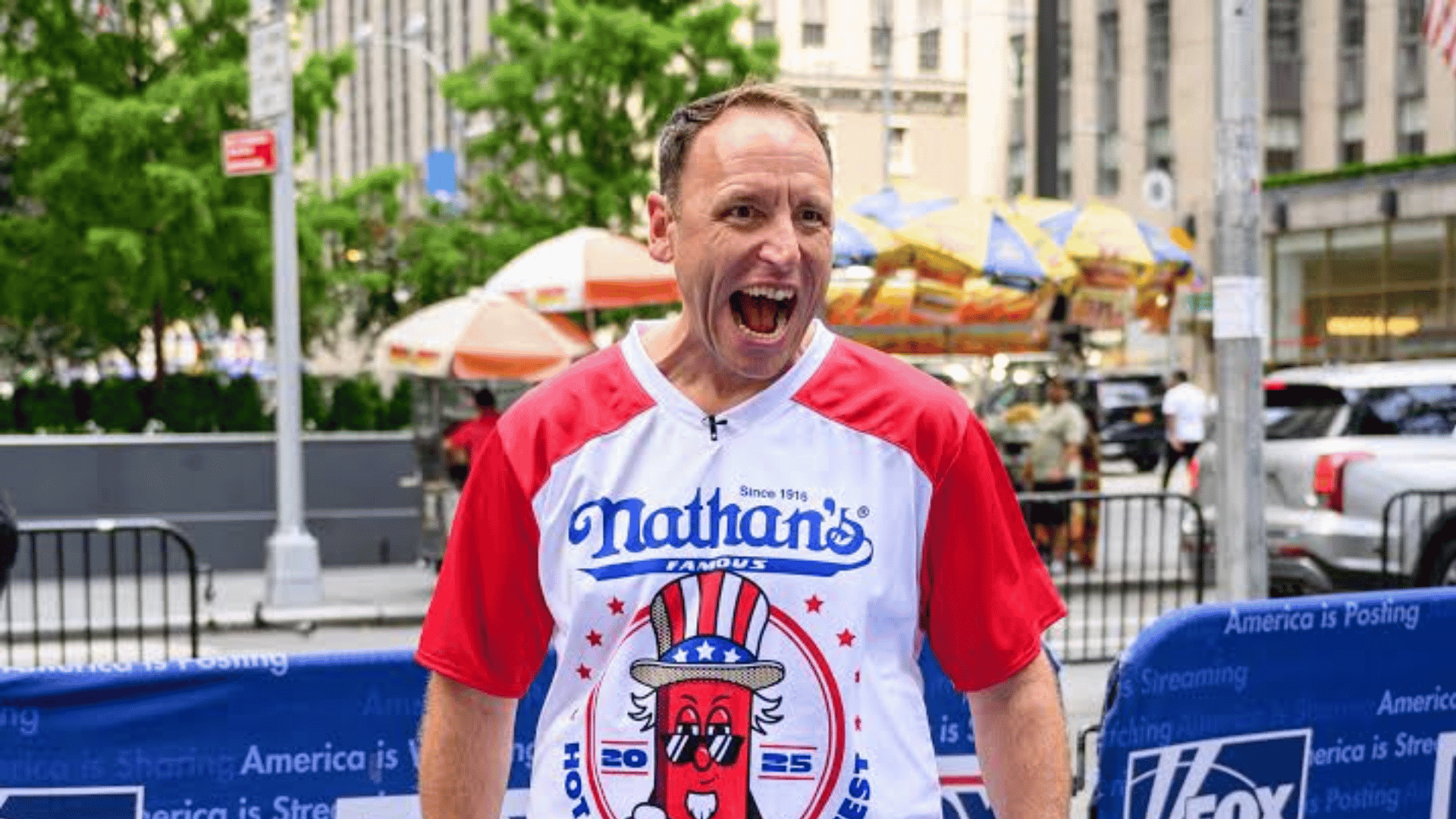 Joey Chestnut wearing a Nathan’s Famous Hot Dog Eating Contest shirt during a public appearance, smiling and celebrating at an outdoor event.