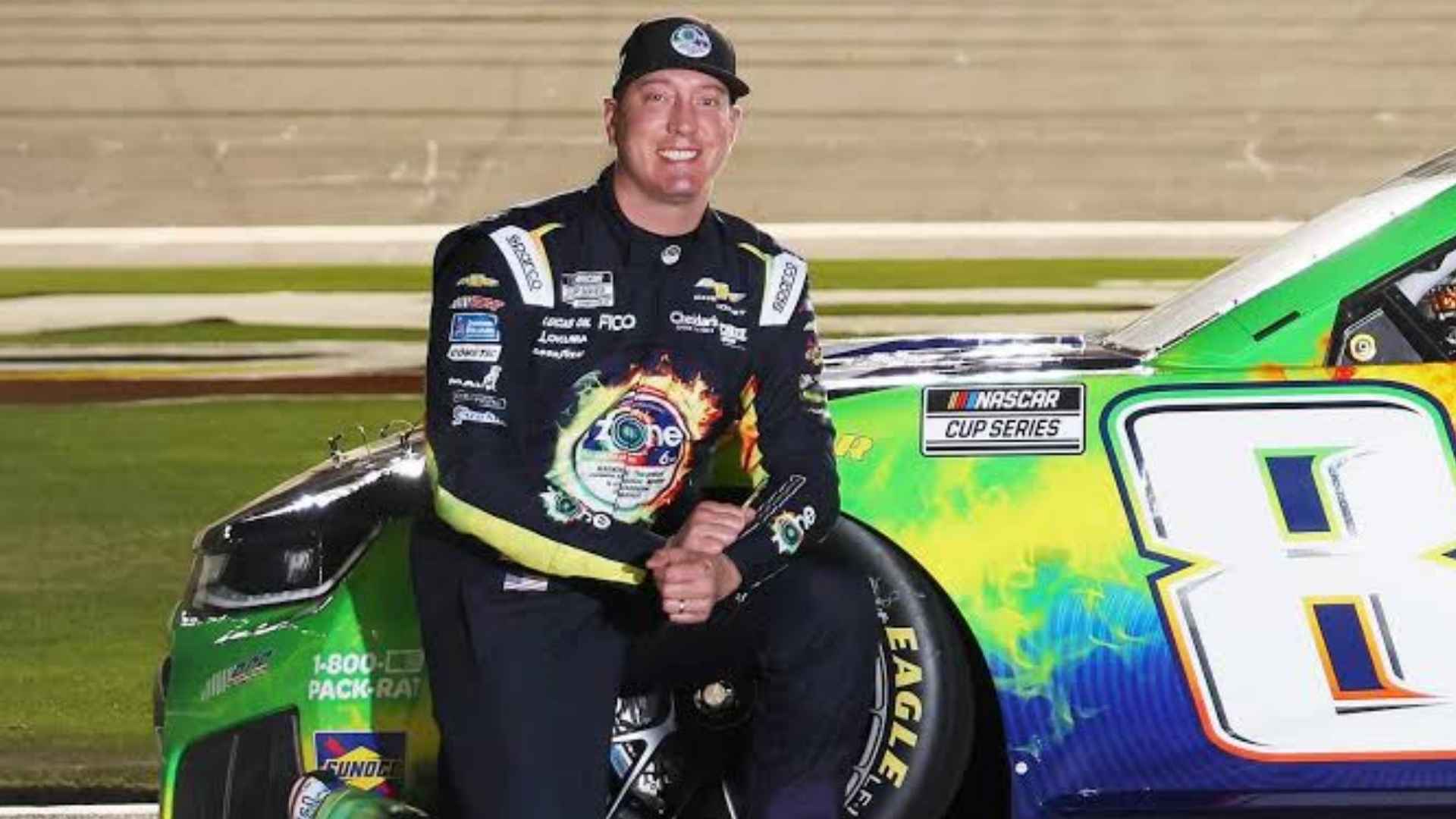 Kyle Busch kneeling next to his green, blue, and yellow No. 8 NASCAR Cup Series race car.