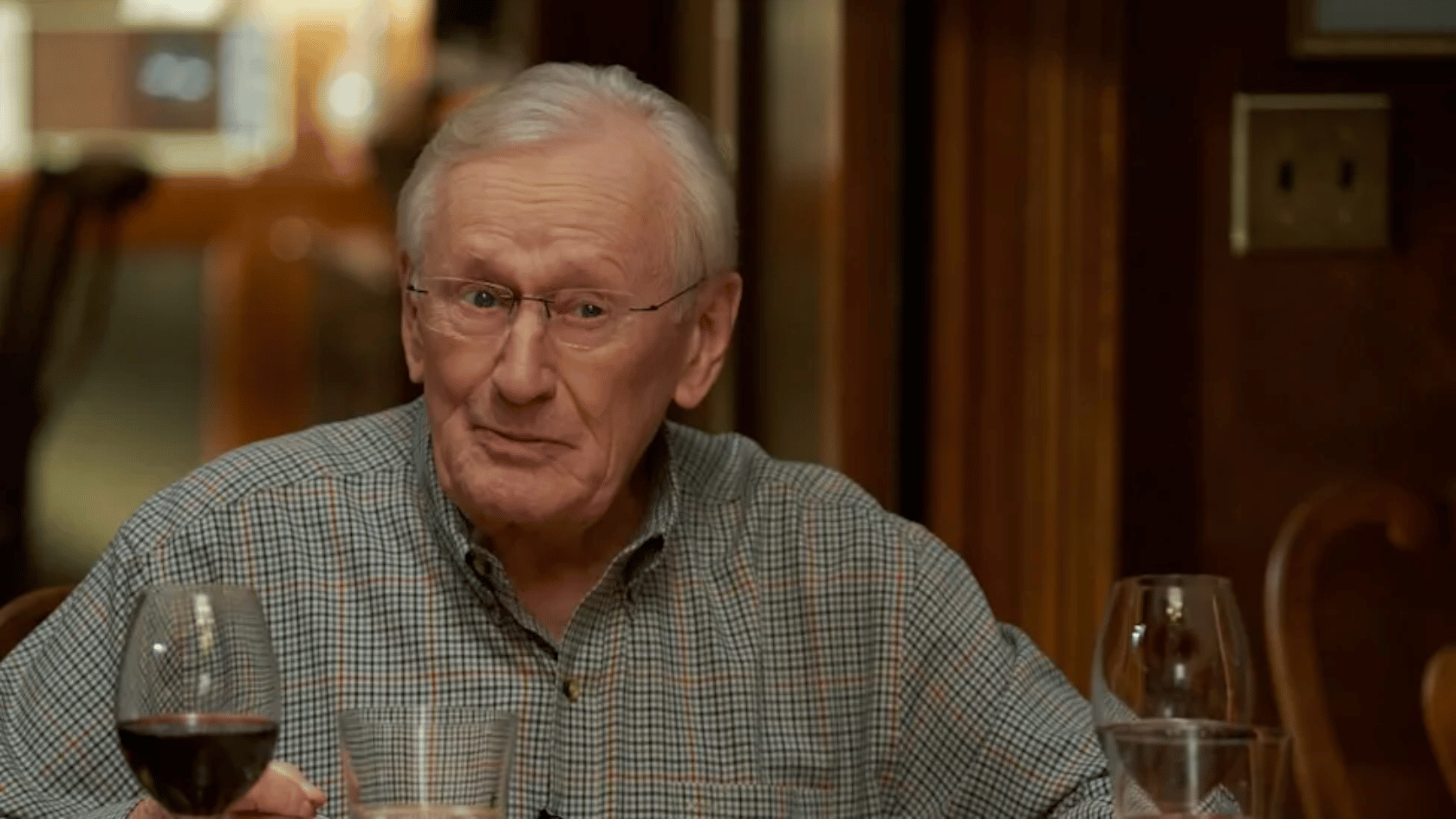 len cariou with white hair and glasses sits at a dining table indoors, wearing a checkered shirt and looking forward with a serious expression