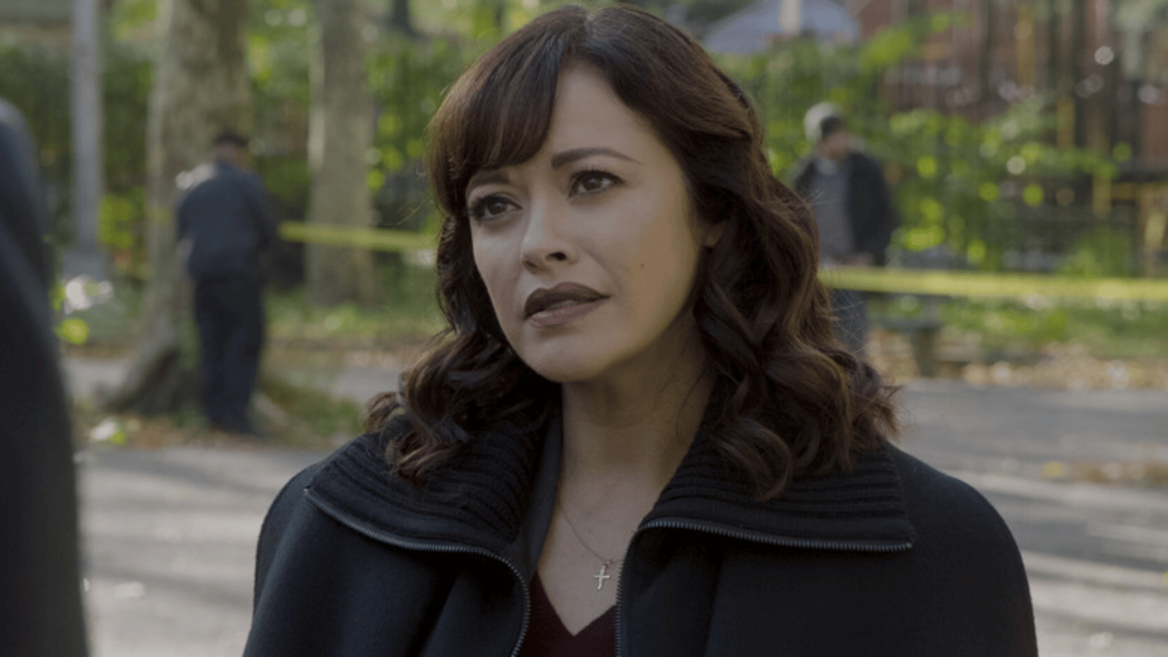 marisa ramirez with dark hair wearing a black coat stands outdoors, looking ahead with a serious expression in a park-like setting