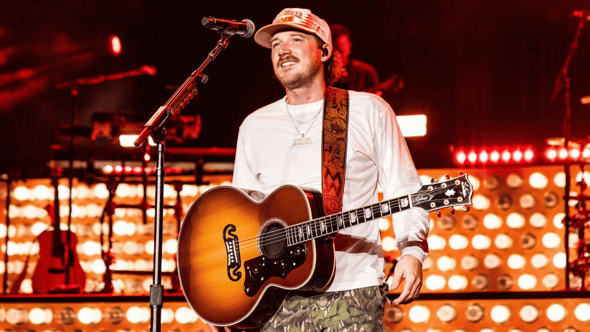 Morgan Wallen performing on stage with an acoustic guitar, singing into a microphone under bright concert lights.