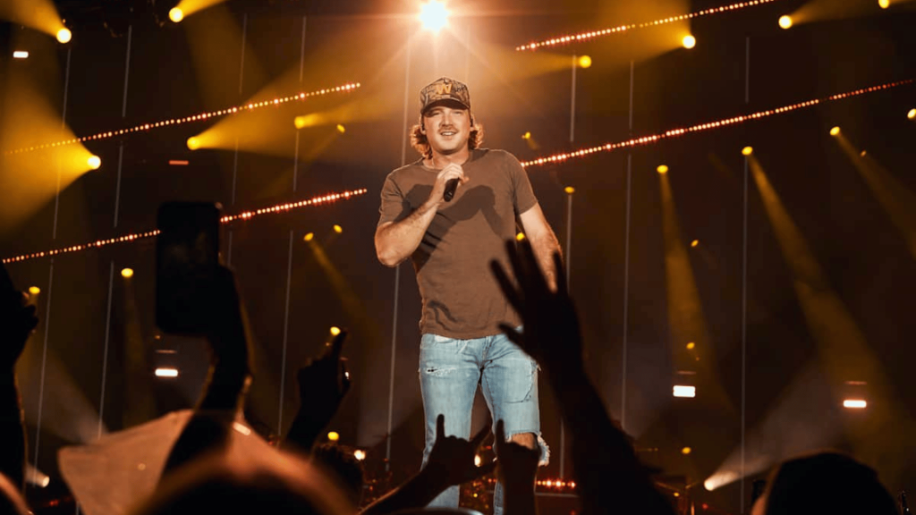 Morgan Wallen performing live on stage with a microphone as fans cheer beneath bright concert lights.