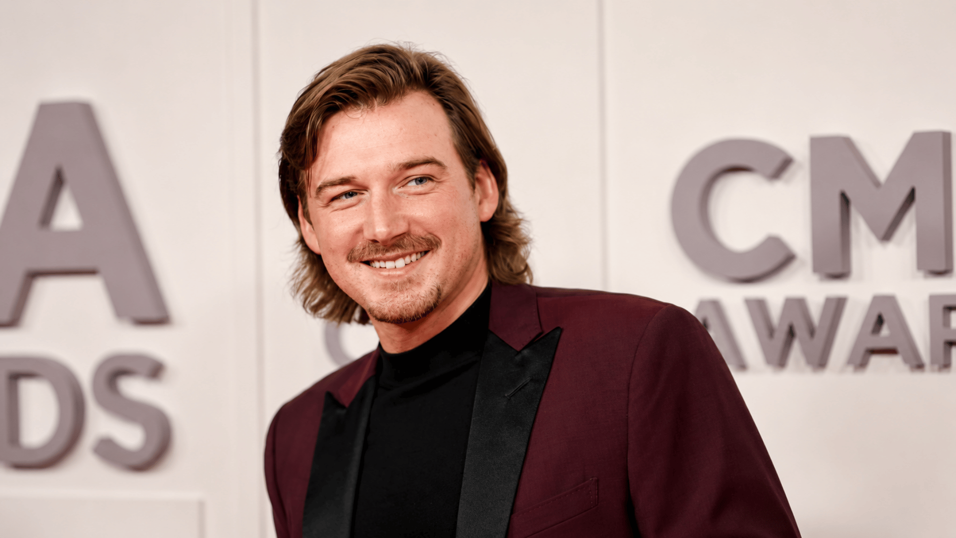 Morgan Wallen smiling on the CMA Awards red carpet while wearing a burgundy suit and black turtleneck.